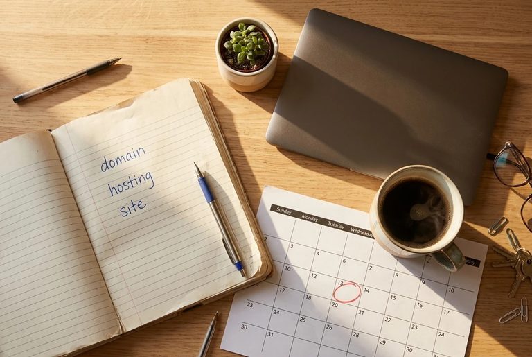 A desk seen from above in warm morning light: a notebook with three handwritten lines for domain, hosting and site, a small potted plant, a coffee mug and a closed laptop, with a p